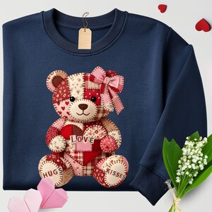 May include: Navy blue sweatshirt featuring a patchwork teddy bear design. The bear holds a heart with "LOVE" on it and has "HUGS" and "KISSES" on its paws. A pink bow adorns the bear's head, perfect for Valentine's Day or a special occasion.