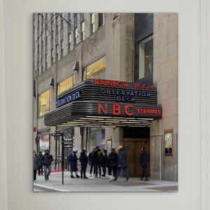 May include: A framed photograph of the NBC Studios entrance, with the words "Rainbow Room" and "Observation Deck" in red neon. A modern gray armchair with a knot-shaped pillow sits in front of the photograph. A small side table with a candle and a silver sphere is next to the chair.