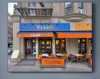 May include: A framed image of a restaurant facade with the name "VIAND" in white lettering. The awning is blue and orange. Tables and chairs are set up outside. The image is hanging on a blue wall, with a white chair and pillows in the foreground.