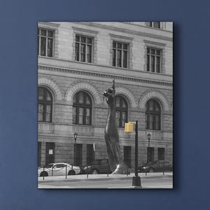 May include: Black and white photograph of a city scene featuring a tall, dark sculpture of a hand pointing upwards. The background includes a large building with multiple windows and parked cars along the street. A yellow traffic light is visible.