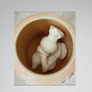 May include: A ceramic chef figurine inside a tan-colored jar. The chef is wearing a white hat and apron. The jar has a wide opening and a slightly textured surface. The image is set against a white wall.