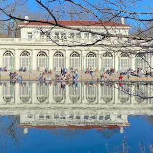 Op de afbeelding: Een wit gebouw met gebogen ramen en een reflecterend zwembad ervoor. Mensen zitten en staan op de trappen van het gebouw.
