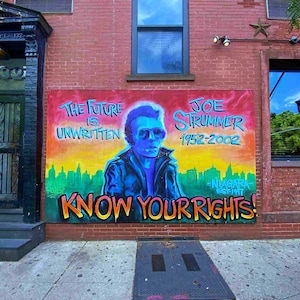 Może przedstawiać: Kolorowy mural na ceglanej ścianie przedstawia Joe Strummera, wokalistę The Clash, z tekstem "The Future is Unwritten" i "Know Your Rights!" Mural zawiera również tekst "Joe Strummer 1952-2002" i "Niagara Esfint".