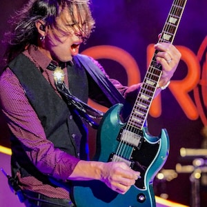 Könnte beinhalten: Eine türkisfarbene E-Gitarre wird auf der Bühne gespielt. Die Person trägt ein weinrotes Hemd mit Knöpfen, eine schwarze Weste und eine Bolo-Krawatte. Der Hintergrund ist dunkel mit Bühnenbeleuchtung.