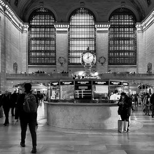 May include: A black and white photo of the interior of Grand Central Station in New York City. The photo shows the main concourse with a large clock in the center and a ticket booth to the right. There are people walking around the concourse.