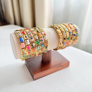 Puede incluir: Pulseras de abalorios doradas exhibidas en un soporte de madera. Cada pulsera presenta numerosos abalorios rectangulares con diseños coloridos, imágenes y texto. Los abalorios están dispuestos en un patrón repetitivo, creando un accesorio visualmente atractivo.