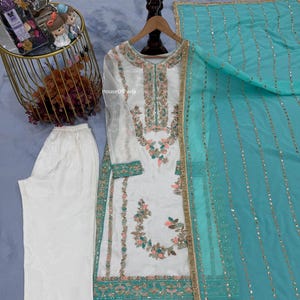 Könnte beinhalten: Weißes besticktes Tunika- und Hosen-Set mit einem türkisfarbenen Dupatta. Die Tunika hat florale Stickereien in Rosa, Grün und Gold. Braune Sandalen und rosa Blumen sind ebenfalls im Bild. Der Text "HouseOfPavla" ist sichtbar.