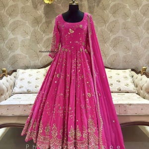 Könnte beinhalten: Ein leuchtend pinkfarbenes, bodenlanges Kleid mit aufwendiger Goldstickerei. Das Kleid hat lange Ärmel, einen fließenden Rock und einen passenden Dupatta. Der Text "Phulkari Shoppe" ist unten im Bild zu sehen.
