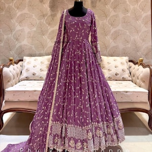 May include: A long, flowing purple dress with intricate gold embroidery. The dress features a round neckline, long sleeves, and a full skirt. The embroidery includes floral and geometric patterns. A matching scarf is draped over the dress form.