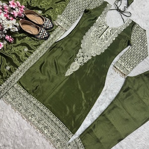 May include: Olive green salwar kameez set with intricate silver embroidery. The set includes a tunic, pants, and a matching scarf. The sleeves and neckline are embellished with detailed mirror work. A pair of matching embellished flats and floral accents complete the ensemble.