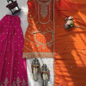 Könnte beinhalten: Ein orangefarbenes, besticktes Kurta-Set mit passendem Dupatta und einem fuchsiafarbenen Rock. Das Kurta hat aufwendige silberne Stickereien. Ein Paar silberne, verzierte Sandalen mit Spiegelakzenten sind ebenfalls vorhanden. Ein kleines Nähmaschinenmodell befindet sich im Hintergrund.