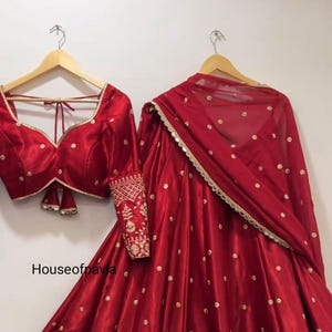Könnte beinhalten: Ein leuchtend rotes indisches Outfit, bestehend aus Oberteil, Rock und Schal, hängt an einem Holzkleiderbügel. Rock und Oberteil sind mit aufwendiger goldener Stickerei und kleinen goldenen Punkten verziert. Das Outfit besteht aus einem glänzenden, seidigen Material.