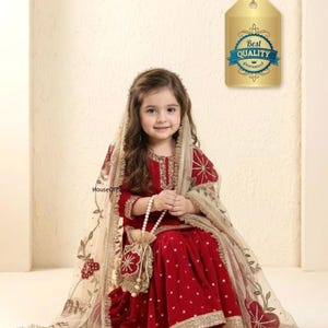 May include: A young girl in a red velvet outfit with gold trim and a matching head covering. The outfit includes a long-sleeved top and a flared skirt. A small beaded purse and a tag that says "Best Quality Guaranteed" are also visible.
