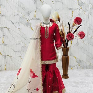 May include: A red and gold embellished salwar kameez outfit with a matching dupatta. The outfit features a long-sleeved top and flared pants. The dupatta is cream-colored with red floral designs and gold trim. The outfit is displayed on a mannequin.