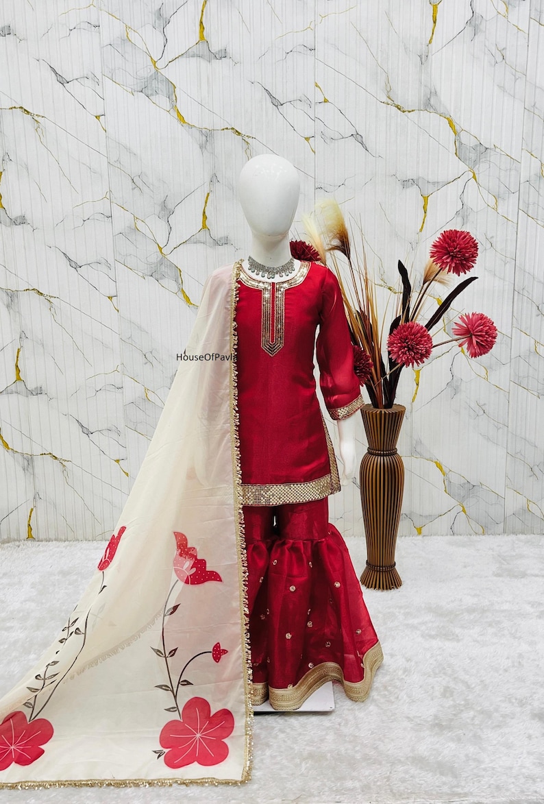 May include: A red and gold embellished salwar kameez outfit with a matching dupatta. The outfit features a long-sleeved top and flared pants. The dupatta is cream-colored with red floral designs and gold trim. The outfit is displayed on a mannequin.