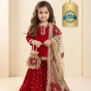 May include: A young girl in a red velvet outfit with gold embroidery and a matching purse. The outfit includes a long-sleeved top, flared pants, and a sheer scarf with floral designs. The outfit has gold trim and a matching purse.