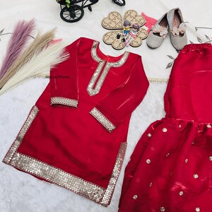 May include: A red, long-sleeved outfit with gold sequin trim. The outfit includes a tunic and a skirt. The tunic has a gold sequin design around the neckline and cuffs. The skirt has gold sequin accents. The outfit is displayed with silver shoes and decorative items.