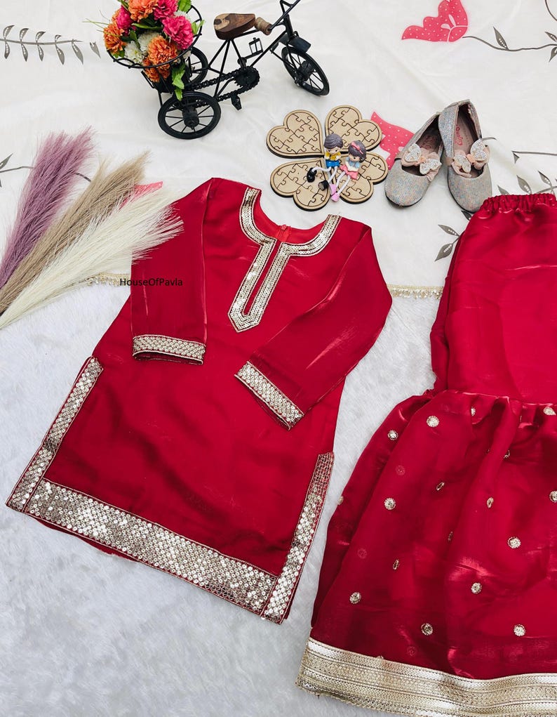 May include: A red, long-sleeved outfit with gold sequin trim. The outfit includes a tunic and a skirt. The tunic has a gold sequin design around the neckline and cuffs. The skirt has gold sequin accents. The outfit is displayed with silver shoes and decorative items.