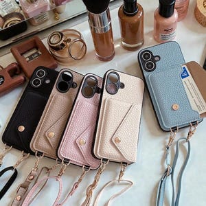 以下が含まれることがあります： 黒、ピンク、ベージュの革製携帯電話ケースのコレクション。各ケースにはカードホルダーと取り外し可能なストラップが付いています。ケースは白い表面に、化粧品やアクセサリーと一緒に展示されています。