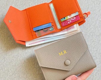 以下が含まれることがあります： 複数のカードスロットとジッパー付きコンパートメントが付いた、開いたオレンジ色の革製財布。現金とカードでいっぱい。イニシャル「M.R」が黄色で入った、閉じた灰色の革製財布が手前にあります。