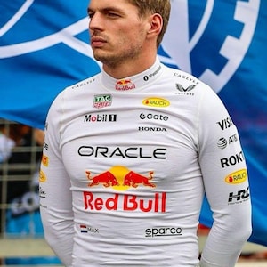 May include: A person wearing a white long-sleeved shirt with various sponsor logos, including "Red Bull" and "Oracle." The shirt features a red and yellow logo on the chest. The person is standing in front of a blue and white flag.