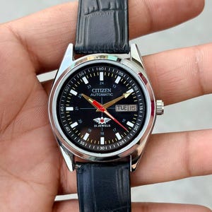 Puede incluir: Un reloj de pulsera automático Citizen con esfera negra y caja plateada. El reloj tiene una correa de cuero negro con un patrón de cocodrilo. La esfera muestra el día y la fecha, con detalles en rojo y dorado. El reloj se sostiene en una mano.