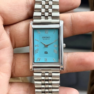 May include: A rectangular Seiko Quartz watch with a light blue face and silver-tone band. The watch face features Roman numerals and the brand name. The band is made of linked metal pieces.
