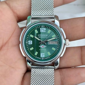 Op de afbeelding: Een automatisch Citizen-polshorloge met een groene wijzerplaat en een zilveren mesh band. De wijzerplaat toont de dag en datum, met de tekst "CITIZEN AUTOMATIC" en "21 JEWELS". Het horloge is gemaakt in Japan.
