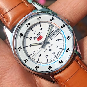 May include: A silver-toned Seiko automatic watch with a white face and brown leather strap. The watch face displays the day and date, with black and blue numerals. The watch has a classic design.