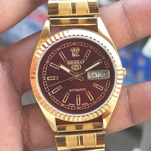 May include: Gold-toned Seiko 5 automatic wristwatch with a burgundy dial. The watch features gold hands and markers, a day-date display, and a gold link bracelet. The dial reads "SEIKO 5 AUTOMATIC JAPAN MADE".