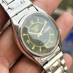May include: A silver Seiko 5 automatic wristwatch with a dark green face. The watch features gold-colored hands and hour markers, a day-date display, and a stainless steel band. The watch face displays the day as "MON 2".