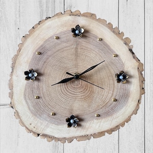 May include: A round wooden clock with a natural wood grain finish. The clock face features black clock hands, brass hour markers, and decorative black flower accents. The clock's diameter is approximately 12 inches.