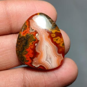 May include: A polished, teardrop-shaped gemstone with a vibrant display of colors. The stone features bands of red, orange, white, and brown, creating a unique pattern. The surface is smooth and reflective, showcasing the natural beauty of the mineral.