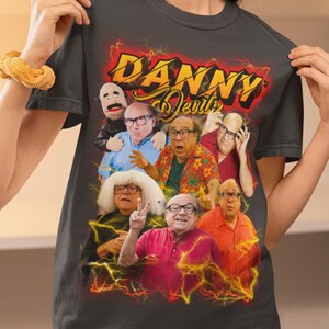 Può includere: T-shirt grigio scuro con una grafica di Danny DeVito in vari ruoli, con la scritta "DANNY Devils" in un design infuocato. Il design include un burattino e diverse immagini di Danny DeVito.