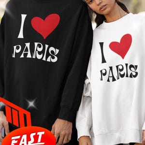 Può includere: Felpe nere e bianche con la scritta "I ❤️ PARIS". Il cuore rosso è sopra la parola "PARIS". Le felpe sono a maniche lunghe e indossate da due persone. L'immagine ha una grafica di consegna rapida.