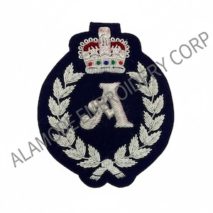 May include: A navy blue embroidered patch featuring a silver crown, the letter "A", and a laurel wreath design. The patch has the text "ALAMO EMBROIDERY CORP" in white. The patch is likely for a uniform or decorative item.