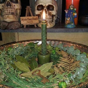 Könnte beinhalten: Eine brennende grüne Kerze in einem Halter, umgeben von Grün und einem antiken Schlüssel in einer dekorativen Metallschale. Zwei Totenkopf-Figuren und andere dekorative Gegenstände befinden sich im Hintergrund. Das Schild lautet "MysticDaisyStudio On Etsy."