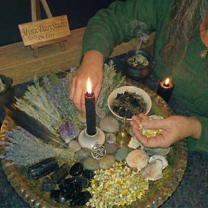 Könnte beinhalten: Eine mystische Anordnung mit einer brennenden schwarzen Kerze, Kräutern, Kristallen und Muscheln auf einem dekorativen Tablett. Das Tablett ist von anderen Kerzen, einer kleinen Glocke und einem Schild mit der Aufschrift "Mystic Daisy Studio On Etsy" umgeben.