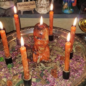 May include: A ritualistic display featuring a central orange figure surrounded by lit orange candles in black holders. The candles are arranged on a silver tray with dried flowers and leaves. A glass of amber liquid is in the background.
