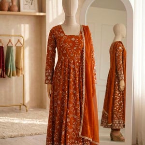 May include: A rust-orange floor-length dress with long sleeves and a square neckline, featuring intricate white floral embroidery and sequin detailing throughout. The garment includes a matching sheer rust-orange dupatta draped over one shoulder.