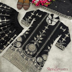 May include: Black embroidered Indian outfit with silver embellishments. The set includes a long-sleeved tunic, matching skirt, and a scarf. The tunic and skirt feature intricate floral and geometric patterns. A pair of embroidered shoes and flowers are also present.