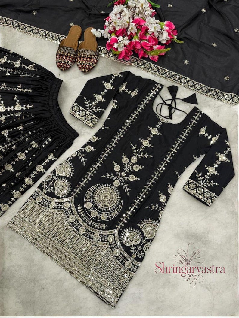 May include: Black embroidered Indian outfit with silver embellishments. The set includes a long-sleeved tunic, matching skirt, and a scarf. The tunic and skirt feature intricate floral and geometric patterns. A pair of embroidered shoes and flowers are also present.