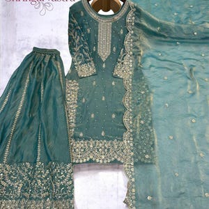 Puede incluir: Un conjunto bordado de tres piezas de color verde azulado. El conjunto incluye una túnica de manga larga, pantalones de pernera ancha y una bufanda a juego. La túnica y los pantalones presentan intrincados bordados y adornos dorados. La bufanda tiene un borde festoneado.