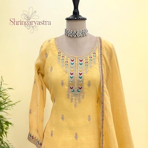 May include: A yellow embroidered salwar kameez outfit on a mannequin. The outfit features intricate embroidery in various colors around the neckline and sleeves. The text "Shringarvastra" is visible on the left side of the image. A silver choker necklace is around the mannequin's neck.