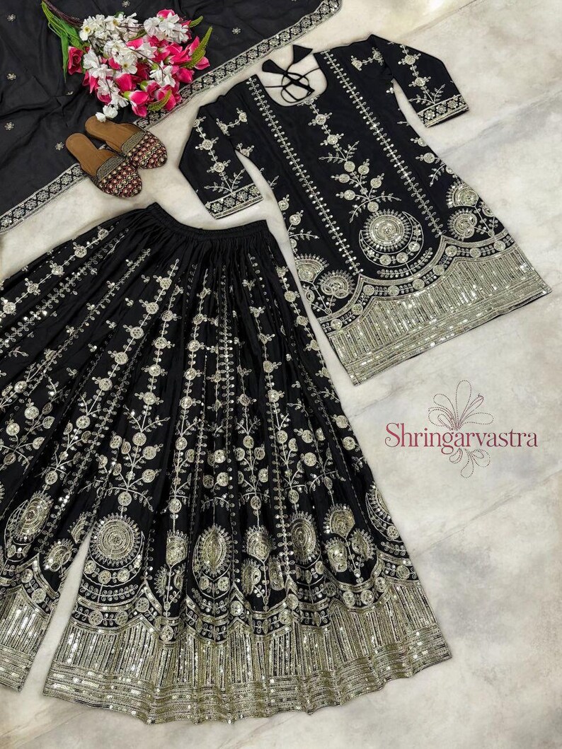May include: A black embroidered three-piece outfit with a matching top, wide-leg pants, and a scarf. The set features intricate silver embroidery and embellishments. A pair of embellished sandals and a bouquet of flowers are also present.