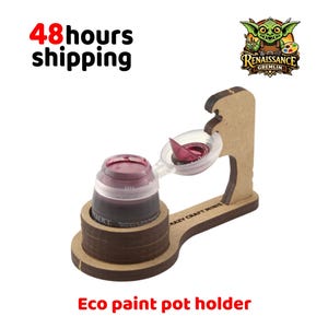 Peut inclure: Un support de pot de peinture en bois marron avec une base circulaire et un bras de support vertical. Le support maintient un petit pot de peinture en plastique transparent rempli de peinture rouge foncé. Le texte "CRAZY CRAFT MINIS" est imprimé sur la base en bois.