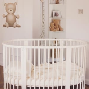 Puede incluir: Una cuna blanca de forma ovalada con barrotes verticales. La cuna está llena de ropa de cama color crema. Una calcomanía de pared de oso de peluche y una decoración de pared con forma de jirafa cuelgan de la pared. Estantes exhiben osos de peluche y fotos enmarcadas.