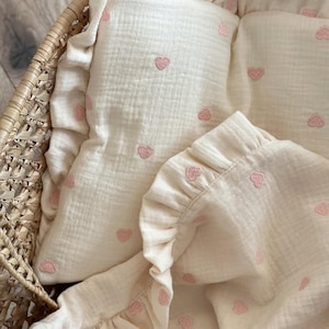 Può includere: Set di biancheria da letto per bambini color crema con cuori rosa ricamati. Il set include un cuscino e una coperta con bordi a balze, il tutto in un cestino di vimini. Il tessuto ha un aspetto strutturato e stropicciato.