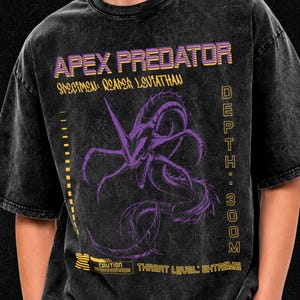 Può includere: Una t-shirt nera lavata all'acido con una grafica viola di una creatura Reaper Leviathan. Il testo include APEX PREDATOR, SPECIMEN: REAPER LEVIATHAN, DEPTH: 300M, CAUTION e THREAT LEVEL: EXTREME in lettere gialle e viola sul davanti della maglia.