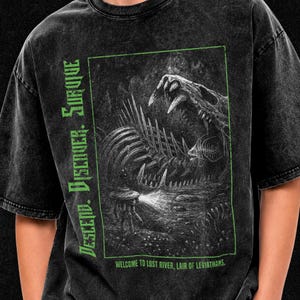 Può includere: Una t-shirt nera con lavaggio acido caratterizzata da una grafica rettangolare verde di una creatura scheletrica in un ambiente buio. Il testo recita "DESCEND. DISCOVER. SURVIVE." verticalmente e "WELCOME TO LOST RIVER, LAIR OF LEVIATHANS." in basso.